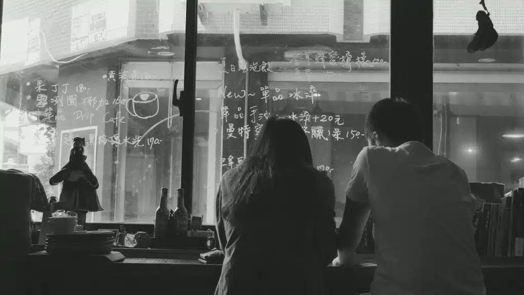 Black and white image of a couple sitting with their backs to the camera, looking out a window at a counter with writing on the glass.