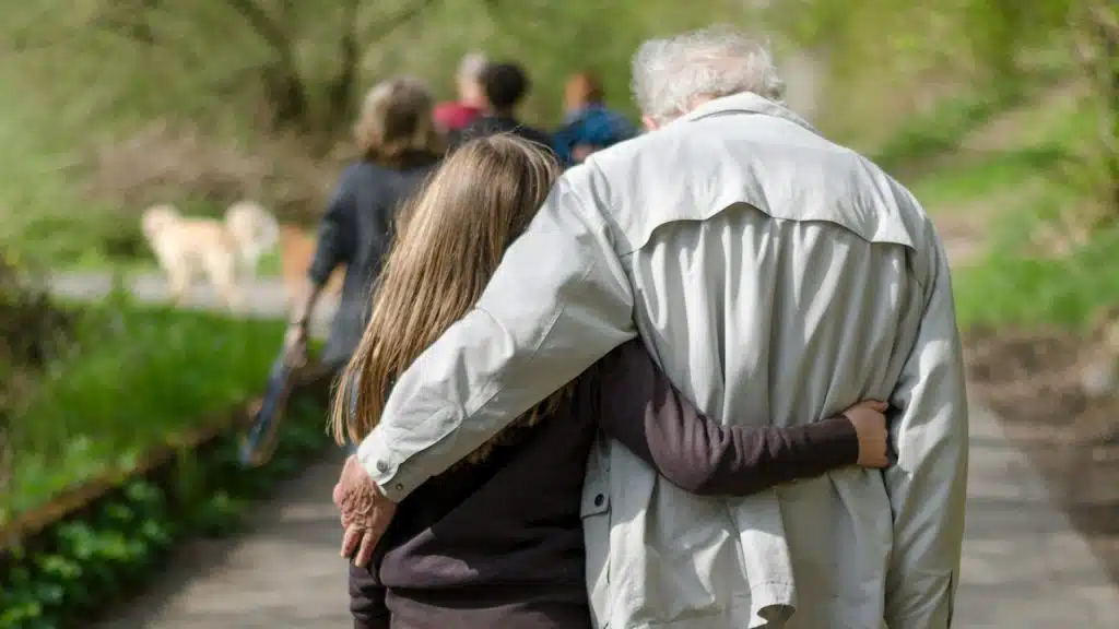 A young woman with an old man walking on a green pathway; the old man is feeling tired.