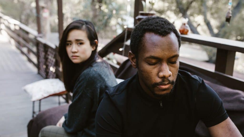 A man with a serious expression and a woman looking upset sitting separately outdoors.