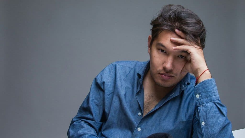 Man in a blue shirt with his head resting on his hand, looking down thoughtfully.