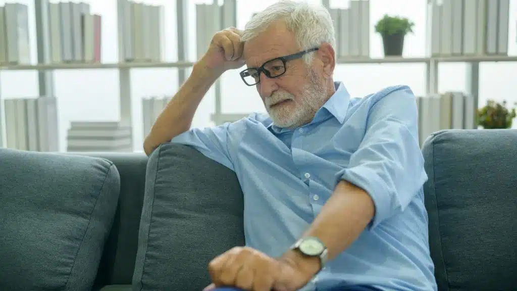 Elderly man in glasses sitting on a gray couch, looking down sadly with a hand on his head.