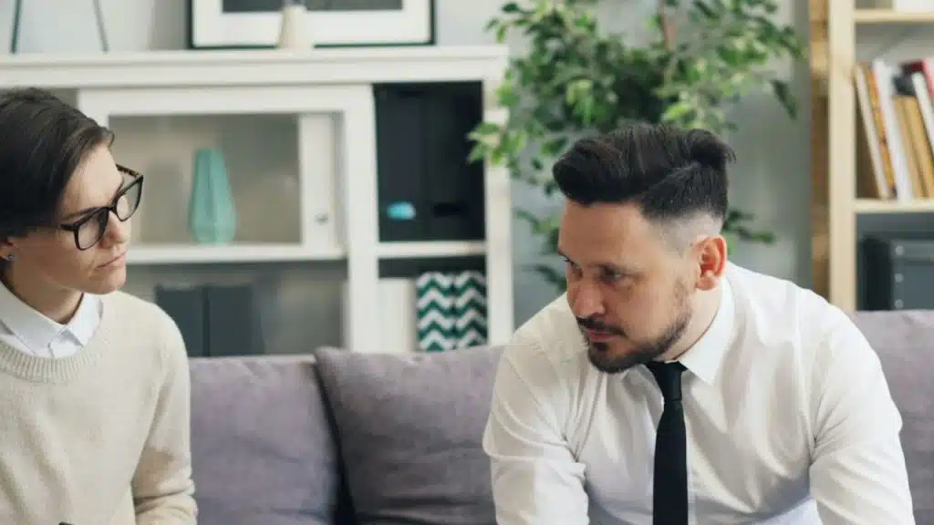Man in white shirt and tie sits opposite a woman with glasses on a couch.