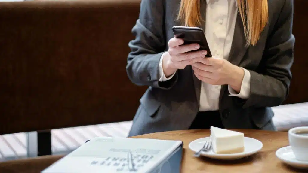 A woman in a suit using her phone at a café table with cake and coffee.