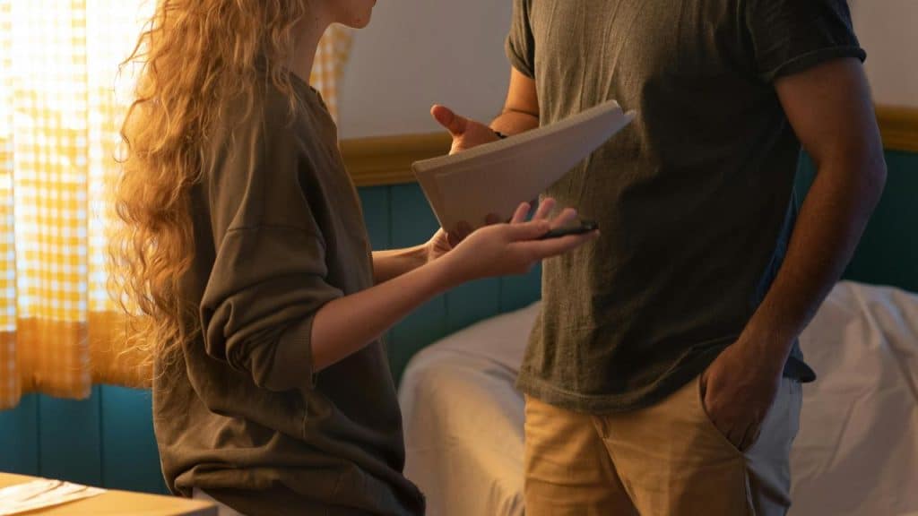 A woman gesturing while talking to a man holding a notebook in a room with moving boxes.