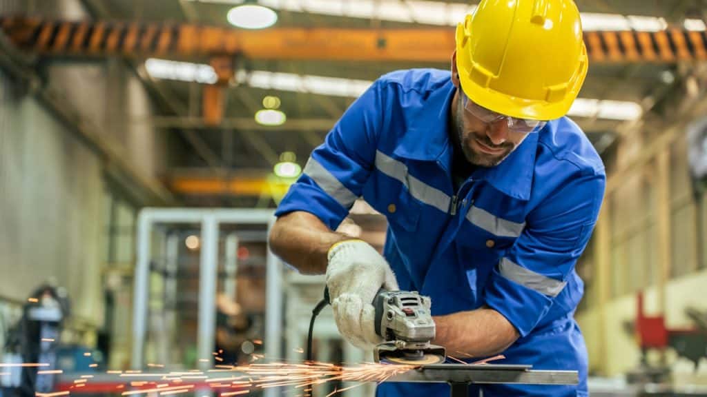 A worker in a blue uniform and yellow hard hat is operating an angle grinder, creating sparks in an industrial setting.