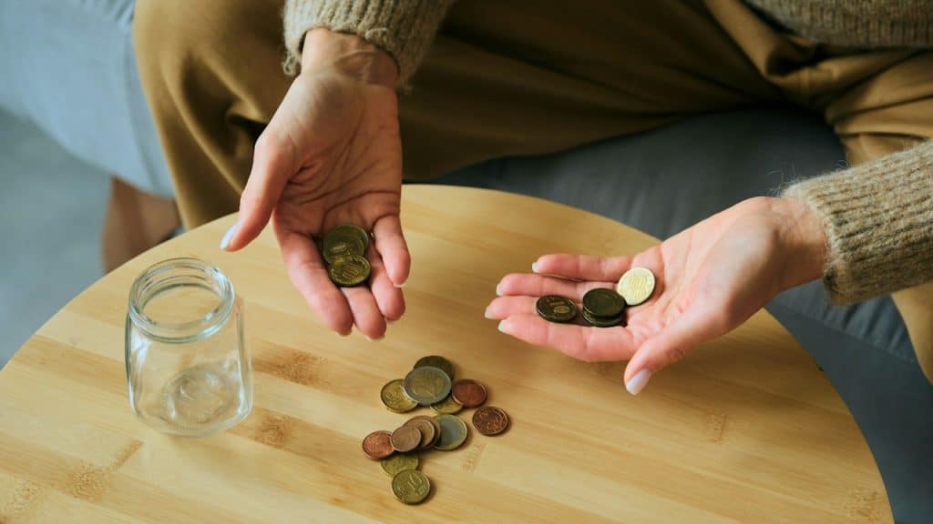 Hands counting coins near a glass jar on a table.