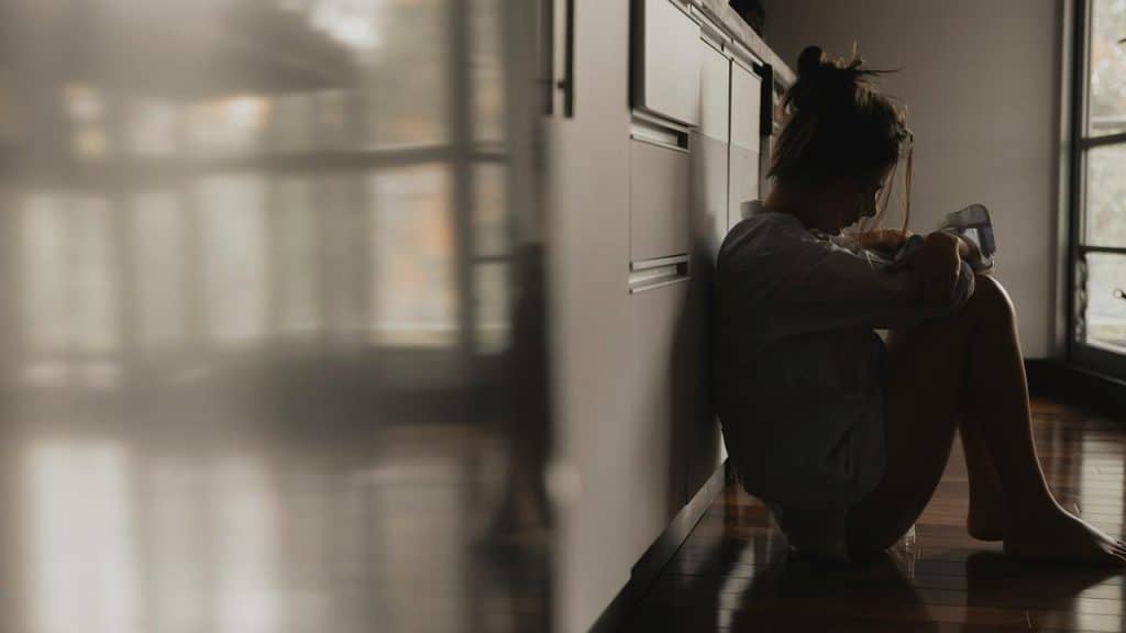 A person sitting on the floor against kitchen cabinets, holding a glass.
