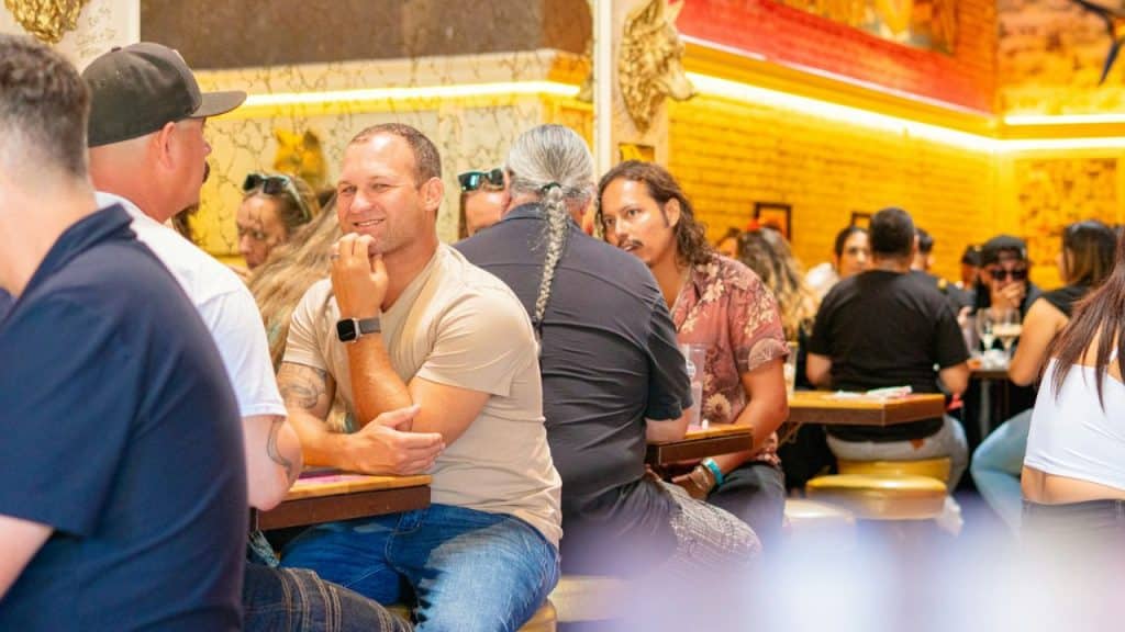 Man in a beige shirt and jeans smiling while sitting at a busy cafe table.