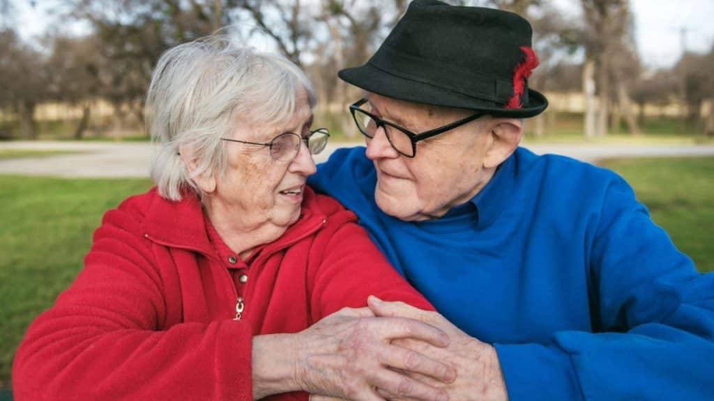 Elderly man and woman with glasses holding hands and looking at each other outdoors.