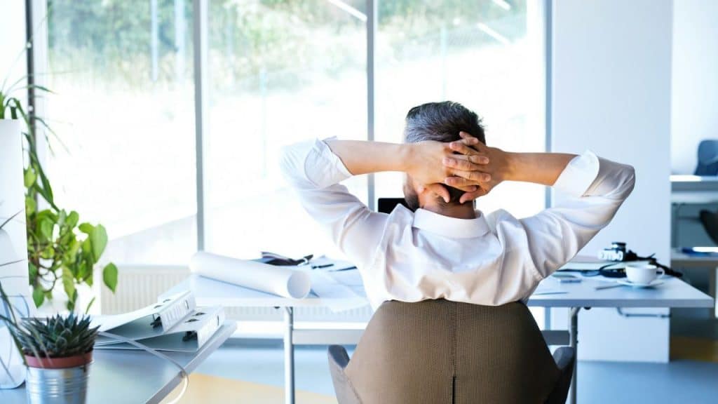 Man with hands behind head relaxes in an office chair, facing away from the camera.