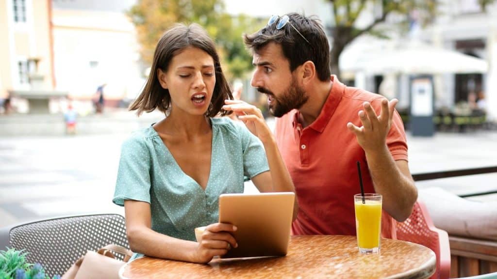 A man in a red shirt gestures and shouts while a woman looks at a tablet.