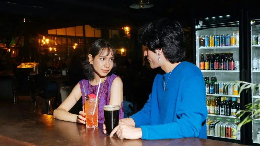 A woman in purple and a man in blue sit at a dark bar with drinks.