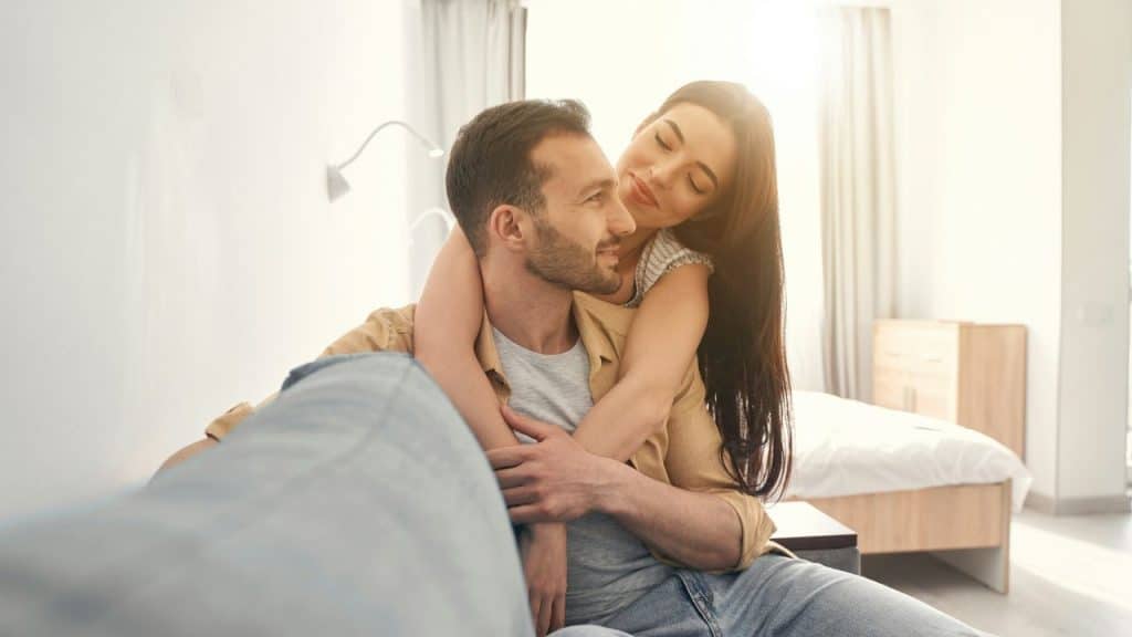 A woman with long dark hair leans over a man's shoulders while they smile at each other.