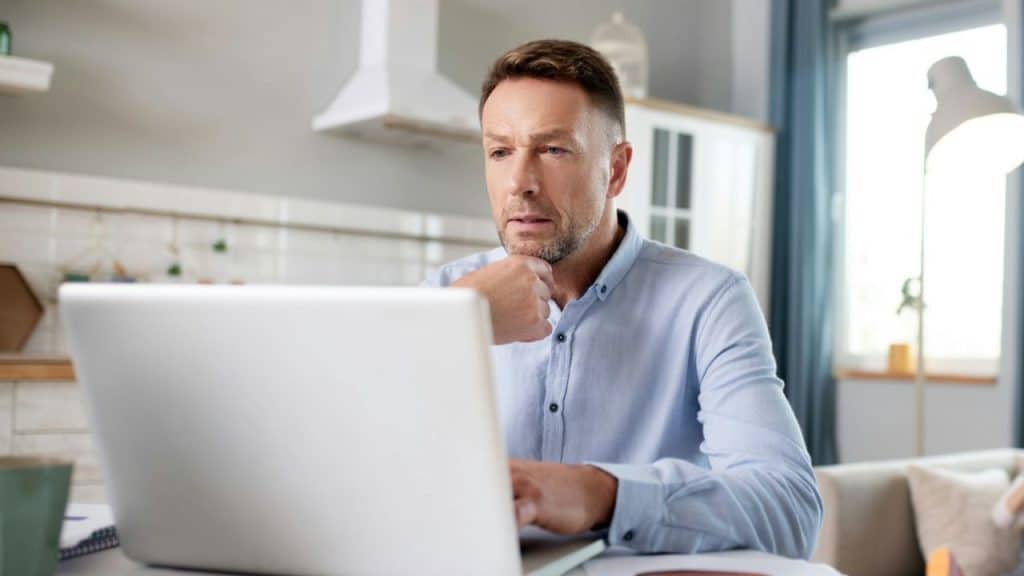 A man in a light blue shirt sits at a table, looking intently at a laptop.