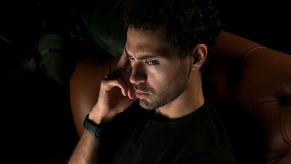 A man in a black t-shirt sits in a dark room, touching his temple.