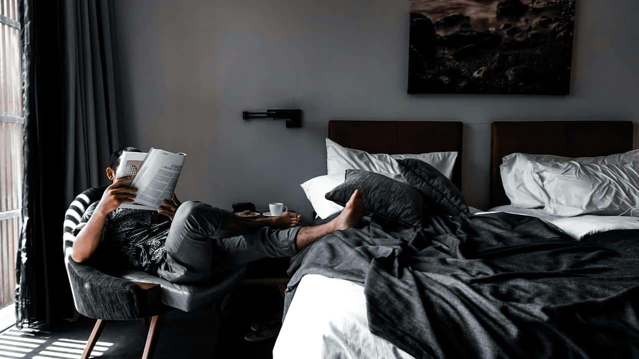 A man reading a book while sitting beside an unmade bed.