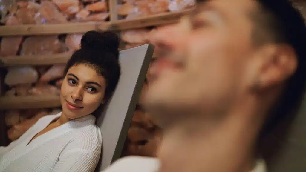 A woman in a white robe smiling while looking at a man relaxing nearby.