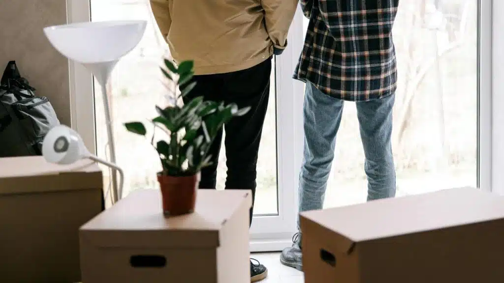 A couple standing by a window surrounded by moving boxes in a bright room.