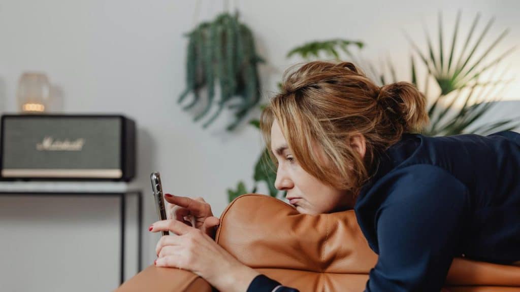 A woman lying on a couch looking at her phone with a sad expression.
