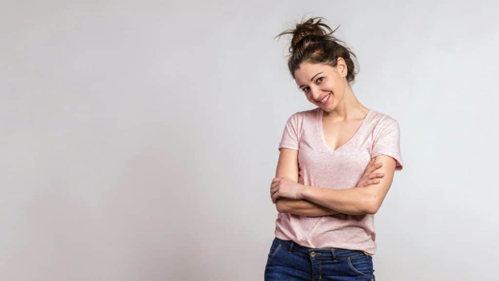 A woman with a messy bun and a pink t-shirt stands with her arms crossed.