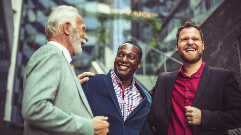 Three men in professional attire stand outside laughing together in front of a building.