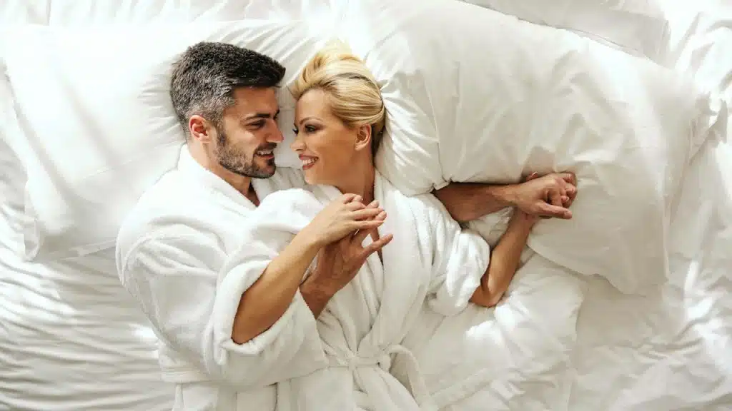 A smiling couple wearing bathrobes and lying in bed while holding each close and gazing in each other’s eyes.