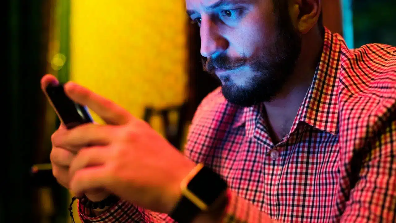 A man in a checkered shirt intensely looking at his phone in colorful lighting.