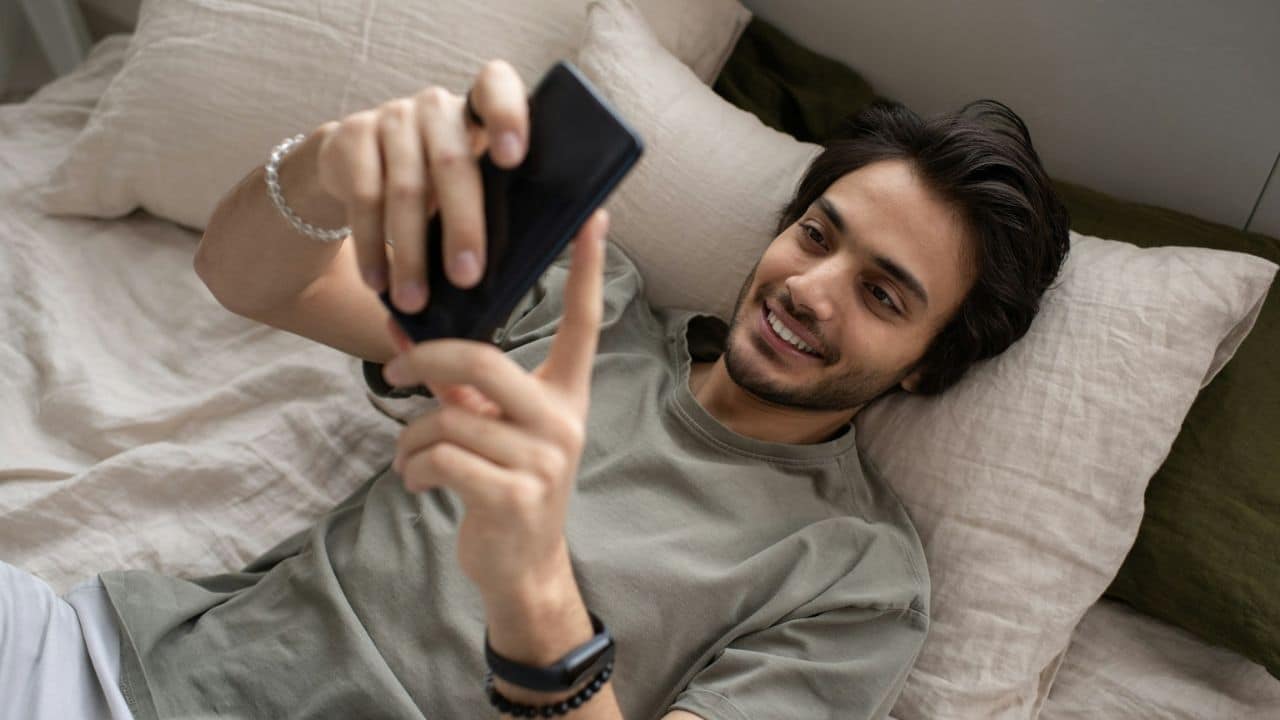 Smiling man lying in bed on pillows, looking at his smartphone.