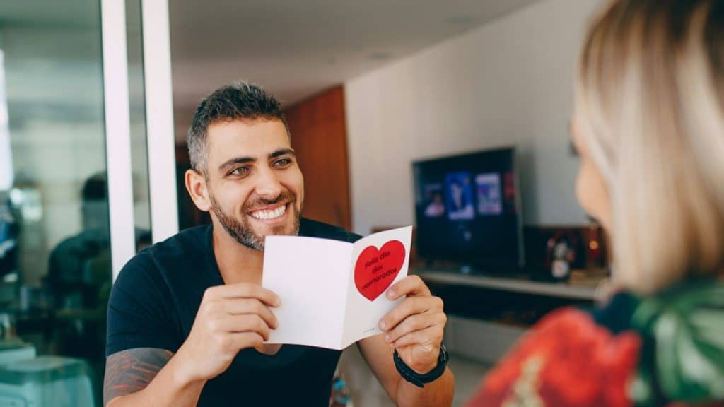 Bearded man in black t-shirt smiles, holding a card with "Feliz dia dos namorados."