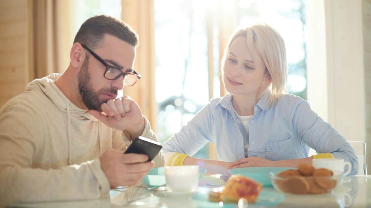 A man looks at his phone at a table while a woman next to him smiles and observes him.