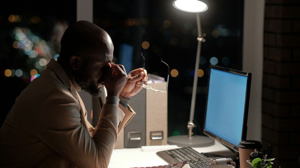 Bald man in a beige jacket rubs his eyes while working at a desk late at night.