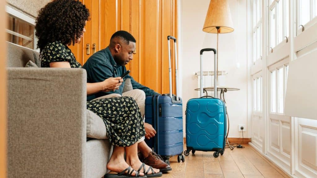 A couple sits on a couch with two blue suitcases, preparing for a trip.