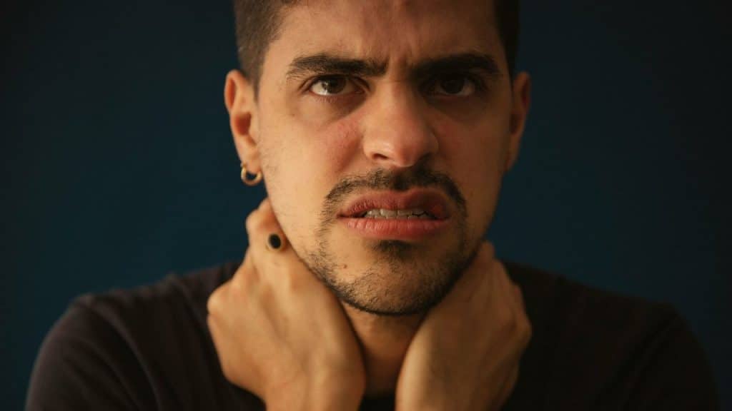A man with a mustache and goatee frowns intensely, holding both hands around his neck.