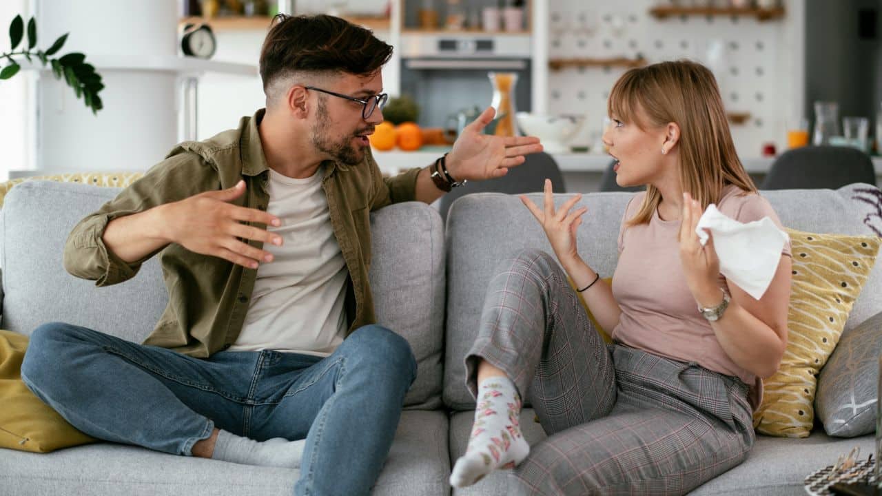 A man and a woman sit on a sofa, gesturing and appearing to argue or discuss intensely.