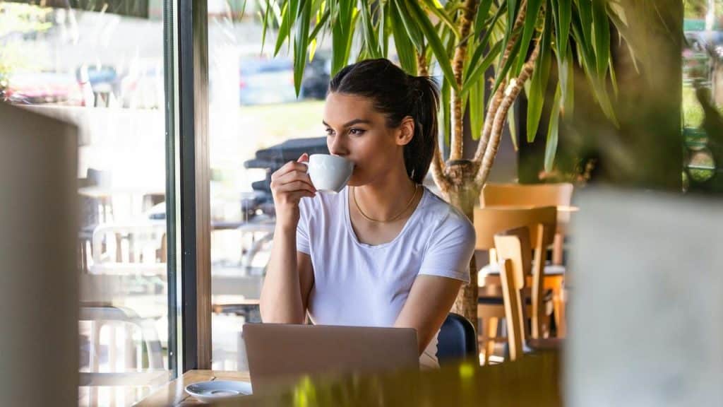 A woman in a white shirt sits at a café table with a laptop, sipping from a cup.
