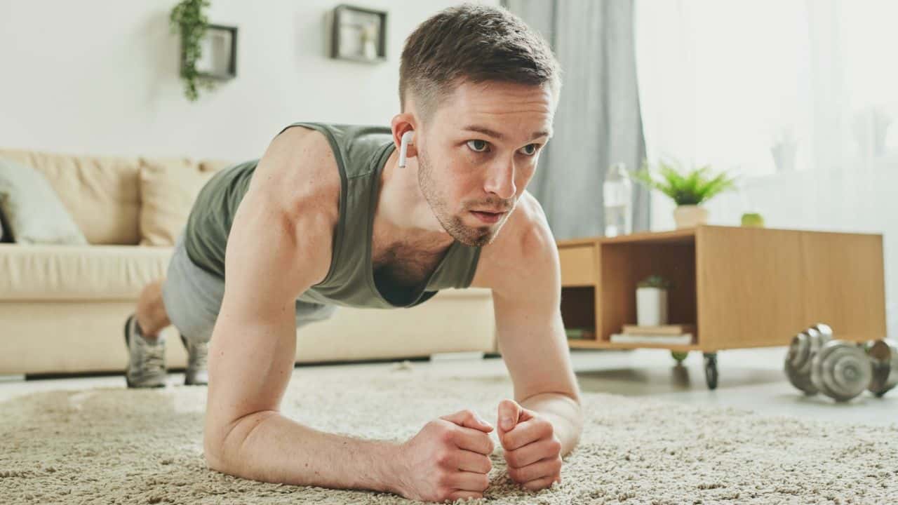 Man in a tank top doing an elbow plank exercise on a rug in a living room.