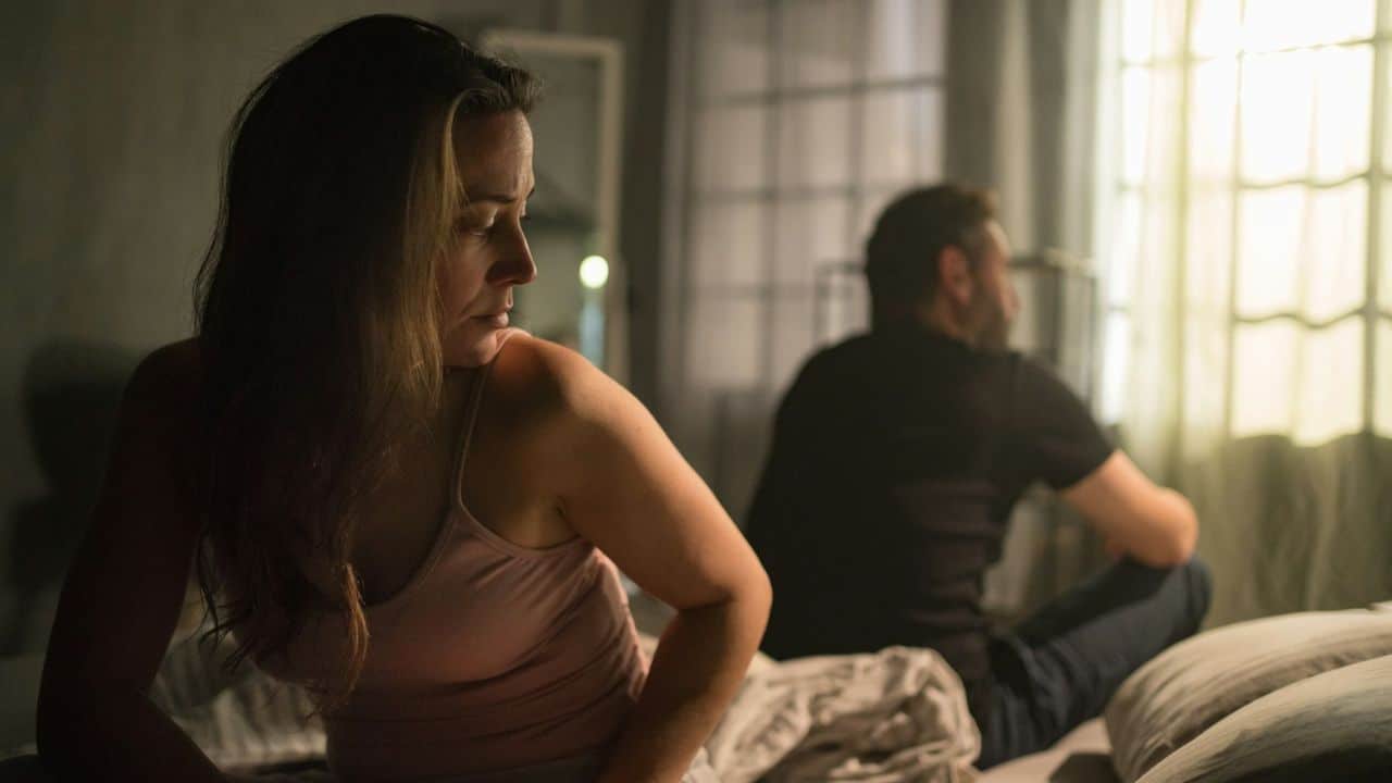 Couple sits back-to-back on a bed in a dim, sunlit room after an argument.