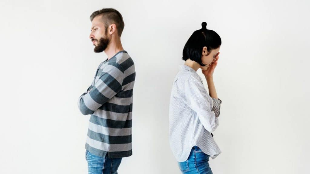 A man and woman stand back-to-back, with the woman holding her head in distress.
