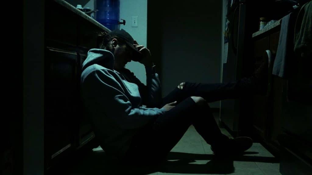 Person in a hoodie and baseball cap sitting on a dark kitchen floor with head in hands.