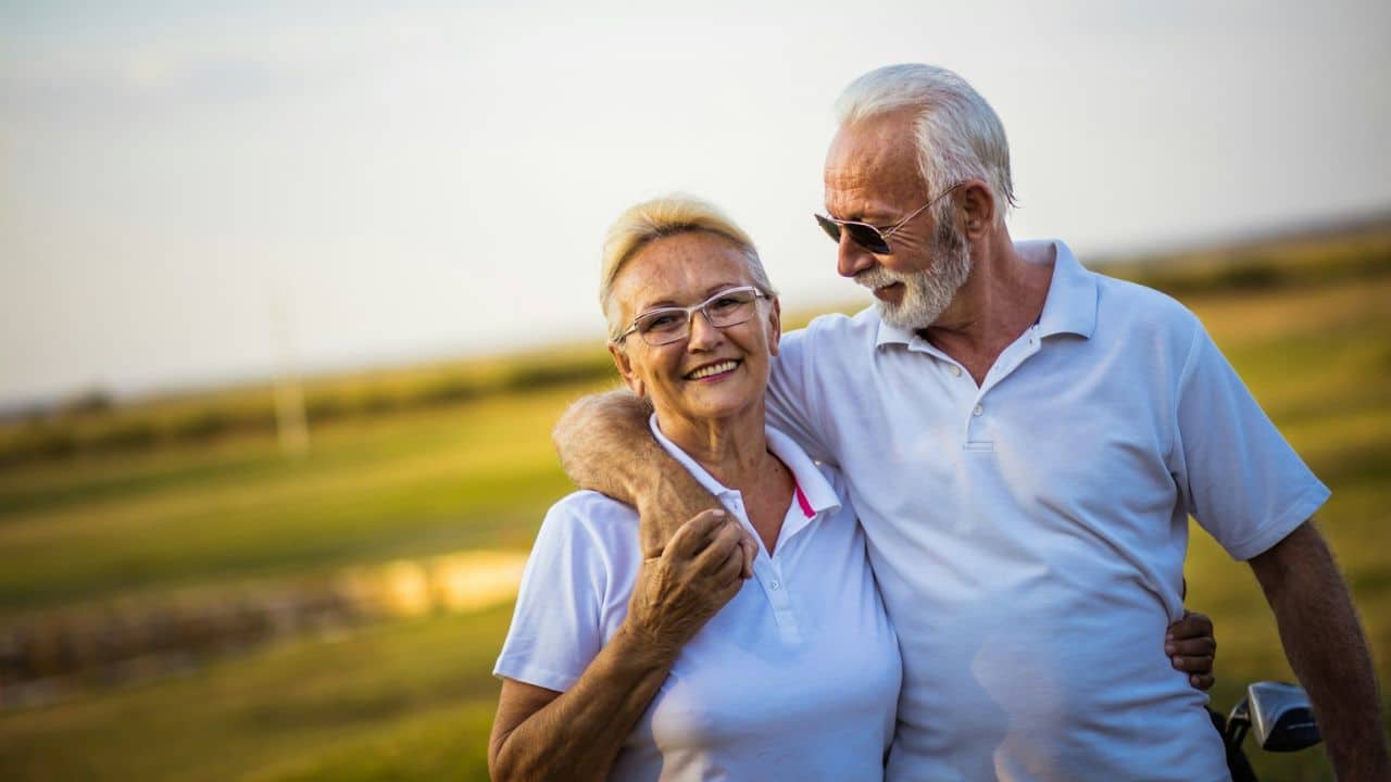 Mature man in sunglasses with his arm around a smiling woman on a grassy field.