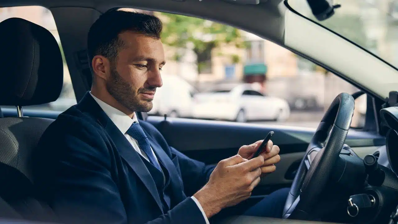 A man in a navy suit and tie is sitting in a car, looking down at and operating his smartphone.