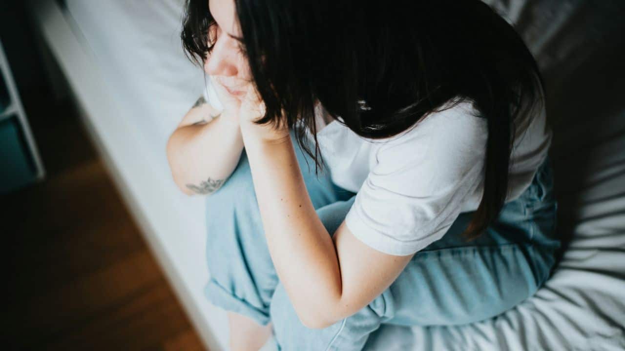 A woman sits on a bed, hugging her knees, looking downcast and covering her face.