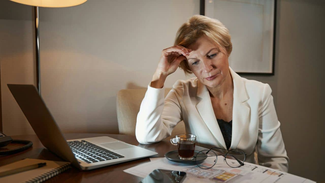 A mature woman looks down at a desk with a laptop and documents.