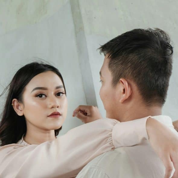A woman looking at the camera with an inscrutable expression while resting her arms on a man’s shoulders.