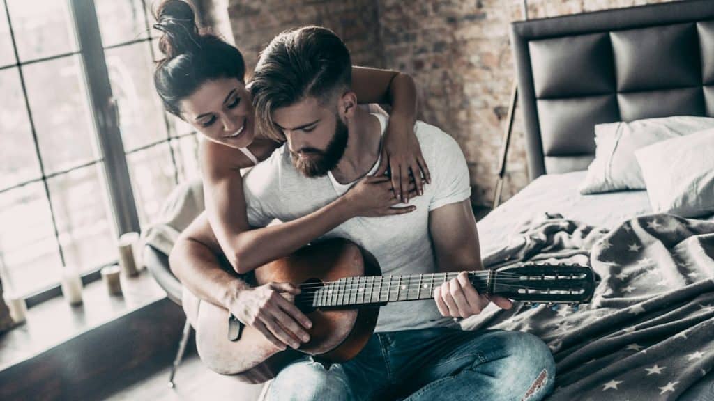 A woman resting her hands on a man’s shoulders whose holding a guitar while they sit on a bed.