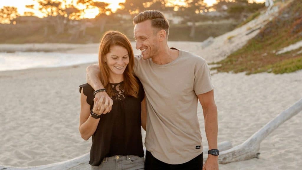 Smiling couple embracing on a beach with a log, with the sun setting behind them.