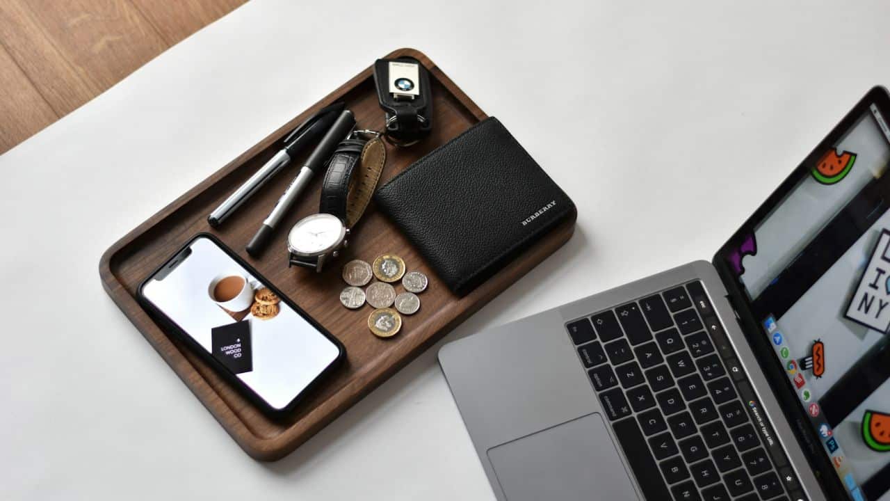 A wooden tray with a wallet, phone, watch, keys, and coins next to a laptop.