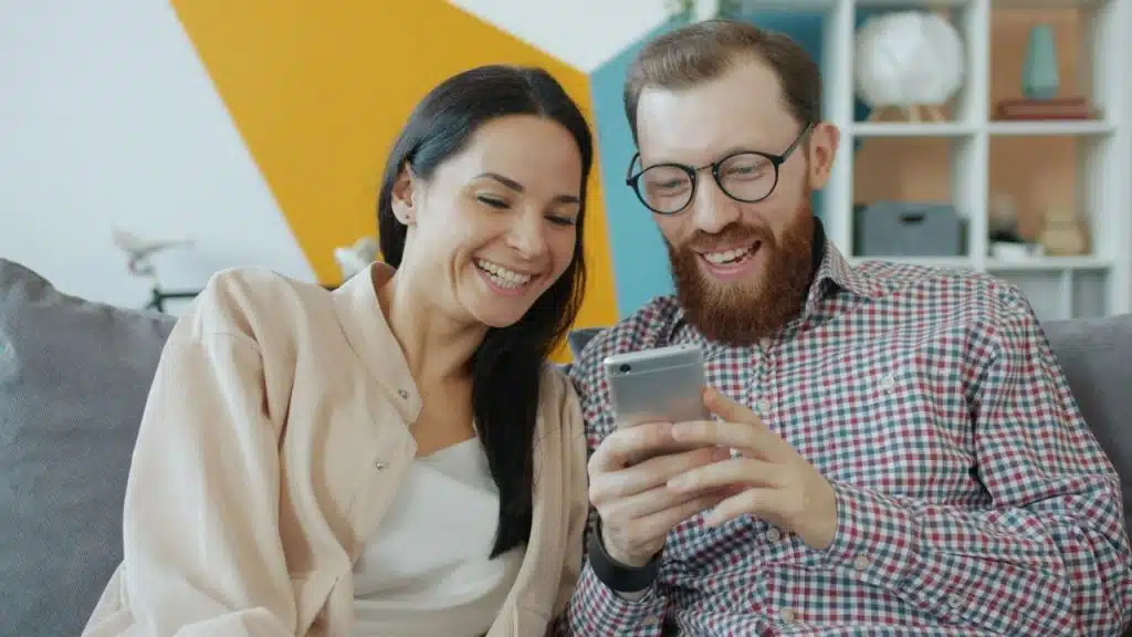 A woman and a bearded man with glasses are laughing while looking at a smartphone.