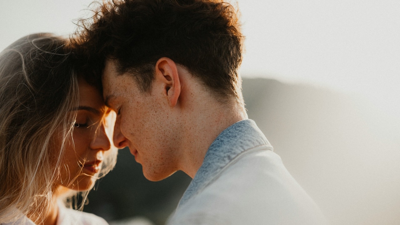 A woman with dark hair is embracing a man with light hair and a beard in a brightly lit room.