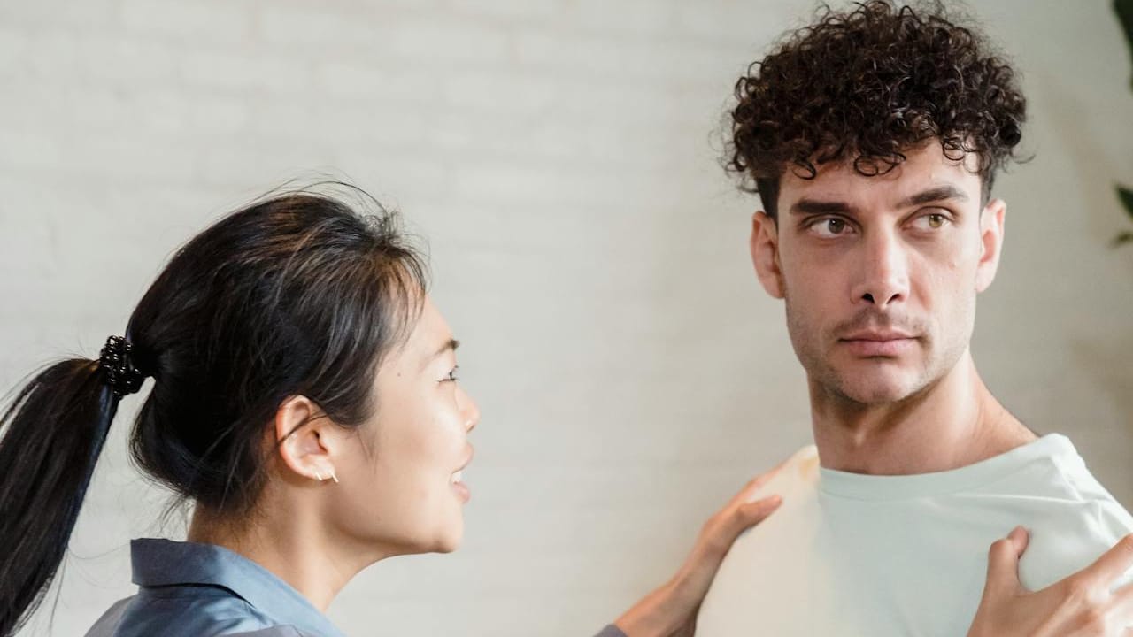 A woman with her hair in a ponytail confronts a man, holding his shoulders as he looks away.